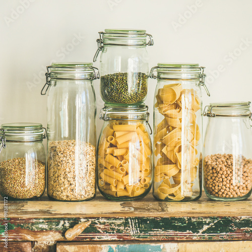 Various uncooked cereals, grains, beans and pasta for healthy cooking in glass jars on wooden table, white background, square crop. Clean eating, vegetarian, vegan, balanced dieting food concept