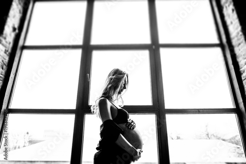 Interesting black and white photo of a beautiful pregnant woman who stands by the big window and sensually hugs her belly