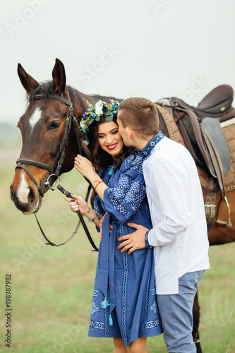 The girl in a beautiful dress and with a wreath on her head holds a horse for a bridle and boyfriend kisses her in the cheek