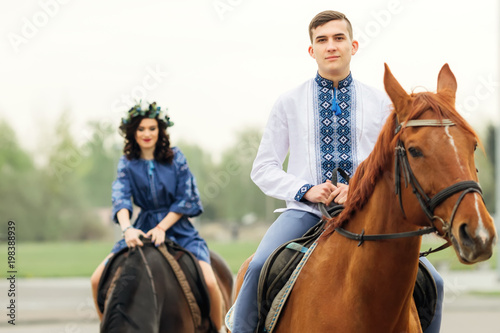 a close-up of a guy in an embroidered shirt who sits on a horse and looks at the camera lens and his girlfriend in the background also sits on a horse © Ivan
