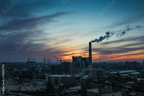 Panorama of city sunset and silhouettes of cranes, high-rise buildings and construction site with smoke