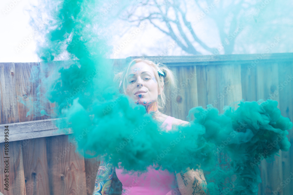 Fototapeta premium Wide portrait of blonde caucasian woman with tattoos using green smoke bomb in an urban location