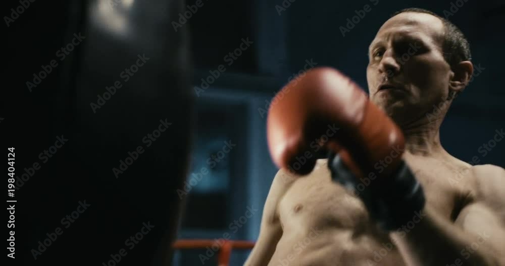 Confident shirtless man training Thai box wearing red gloves and hitting punching bag on ring in gym. Confident shirtless man training Thai box wearing red gloves and hitting punching bag on ring in gym.