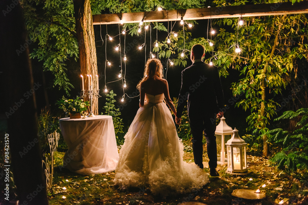 Night wedding ceremony with candles, lanterns and lamps on tree. Bride and  groom holding hands on background of baulb lights, back view. Beautiful  young couple standing under a tree at night Stock