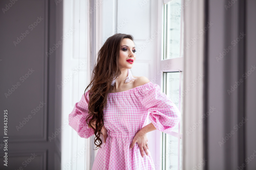 young beautiful girl stands at the big window in the floor and looks out into the distance. Dressed in a light checkered pink dress, long hair, red lips young beautiful girl stands at the big window in the floor and looks out into the distance. Dressed in a light checkered pink dress, long hair, red lips