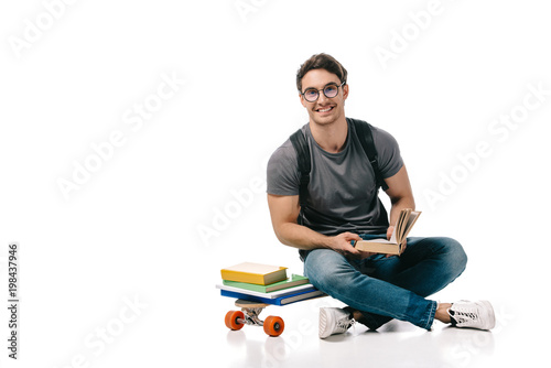 smiling handsome student sitting on skate and holding book on white