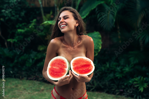 Lovely brunette girl with long curly hair laughing. A beautiful woman is holding watermelons in her hands. Beautiful female body, model covers the bare chest with fruit.
