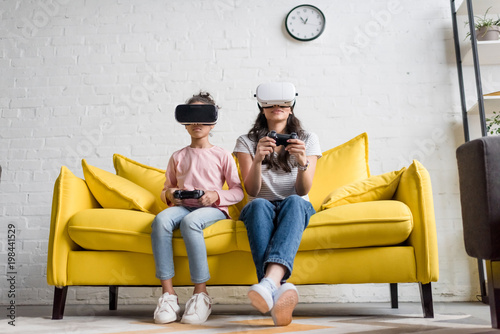 young mother and daughter in vr headsets playing video games at home on couch