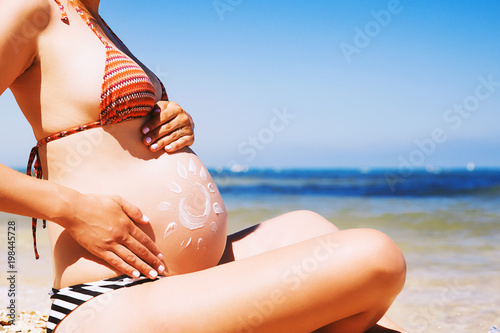 Pregnant woman skin care moisturizer cream on belly on beach at summer