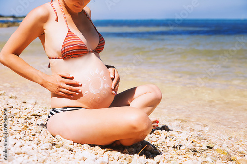Pregnant woman skin care moisturizer cream on belly on beach at summer