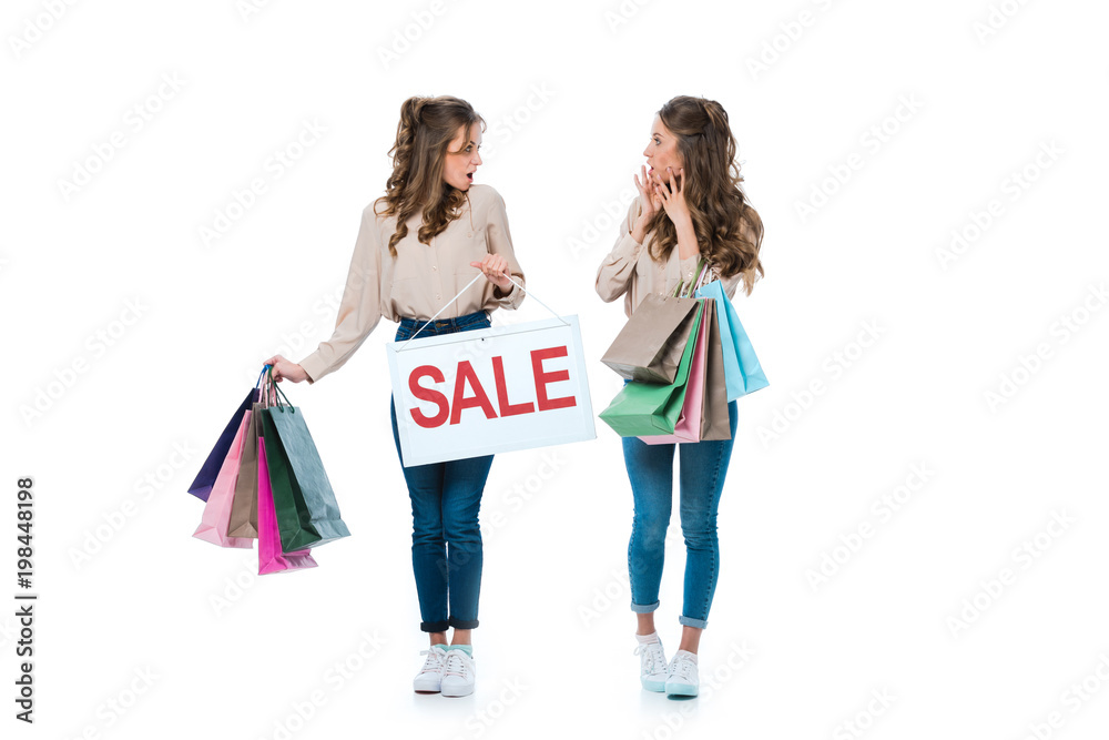 surprised young twins with sale sign and shopping bags isolated on white
