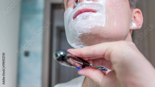 A young man has already shaven half of his beard. There is still a lot of foam on his face.