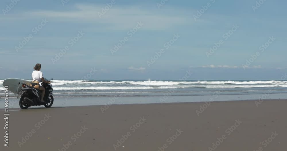 Young attractive girl driving scooters with surfboard on the beach near the water. Shooting in motion from behind Young attractive girl driving scooters with surfboard on the beach near the water. Shooting in motion from behind