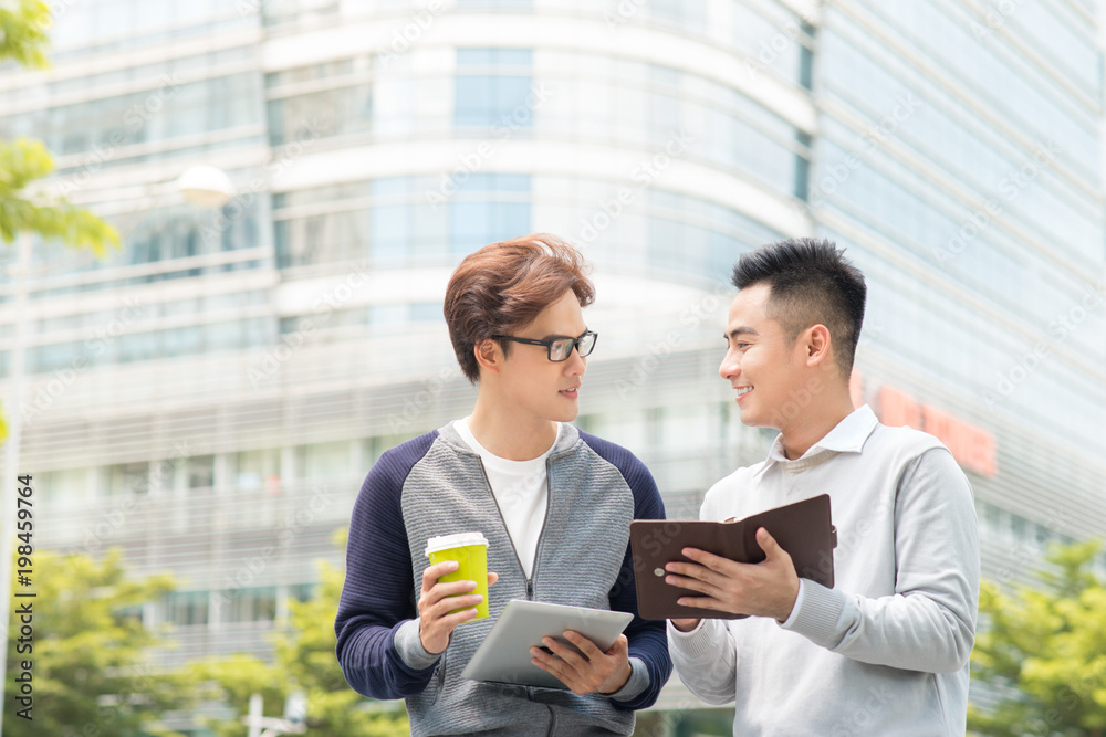 Two smiling male work coworkers outdoors talking together over a digital tablet Two smiling male work coworkers outdoors talking together over a digital tablet