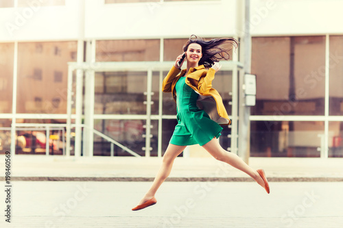 technology, communication and people concept - smiling young woman or girl calling on smartphone on city street and jumping high