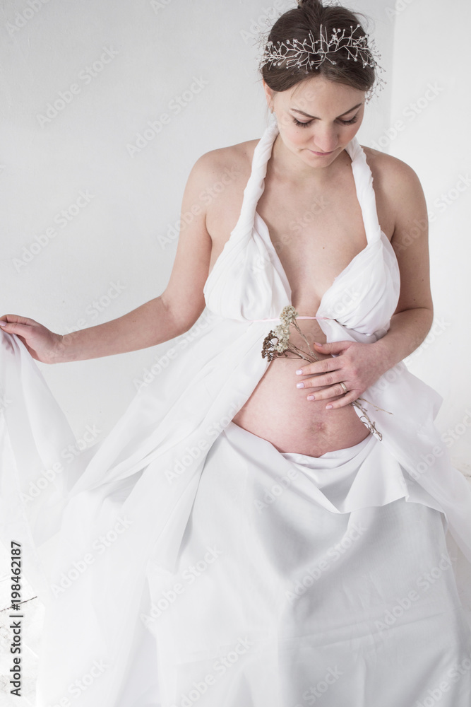 photo Pregnant girl in a diadem and white drapery with flowers in her hands photo Pregnant girl in a diadem and white drapery with flowers in her hands