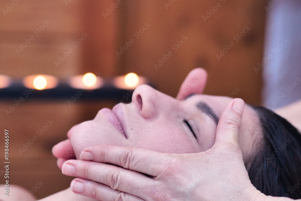 Naklejka premium A beautiful young girl (woman) having a massage in the spa salon, a romantic setting, a pink flower behind the ear. Concept: spa procedures, body massages, spa creams, relax, spa water treatments.