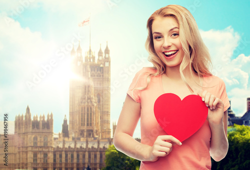 love, travel, tourism, valentines day and people concept - smiling young woman or teenage girl with blank red heart shape over london city background