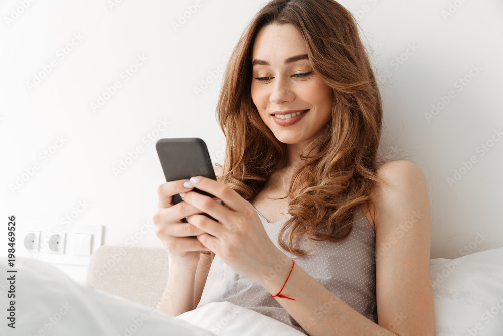 Portrait of young cheerful woman resting in bed after sleep with white clean linen at bedroom, and using smartphone Portrait of young cheerful woman resting in bed after sleep with white clean linen at bedroom, and using smartphone