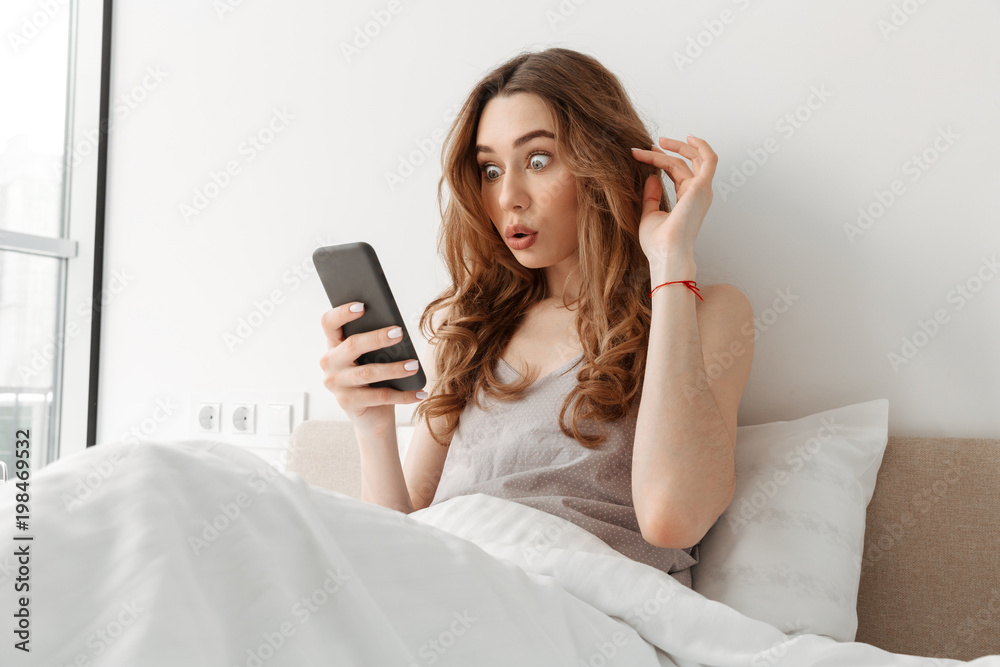 Surprised woman 20s using cell phone, while lying in white cozy bed in morning Surprised woman 20s using cell phone, while lying in white cozy bed in morning