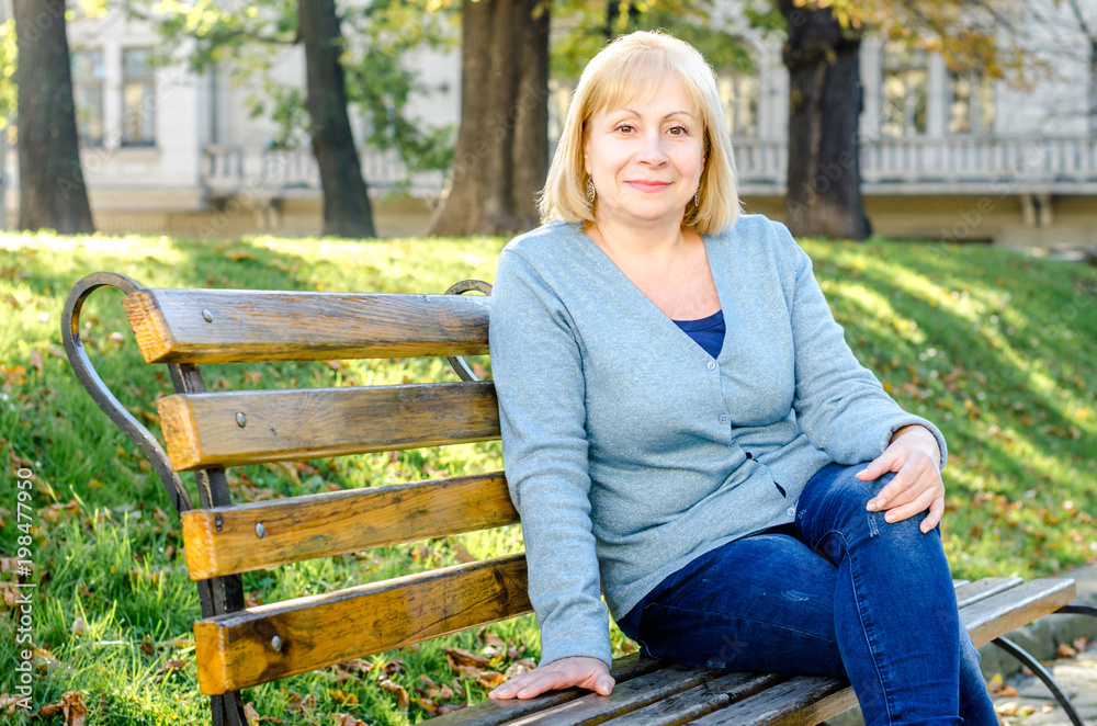 Middle-aged blond woman sitting on a bench in the park looking at the camera on the autumn spring background. Portrait of beautiful senior happy elderly blond woman in a park on a sunny day. Middle-aged blond woman sitting on a bench in the park looking at the camera on the autumn spring background. Portrait of beautiful senior happy elderly blond woman in a park on a sunny day.