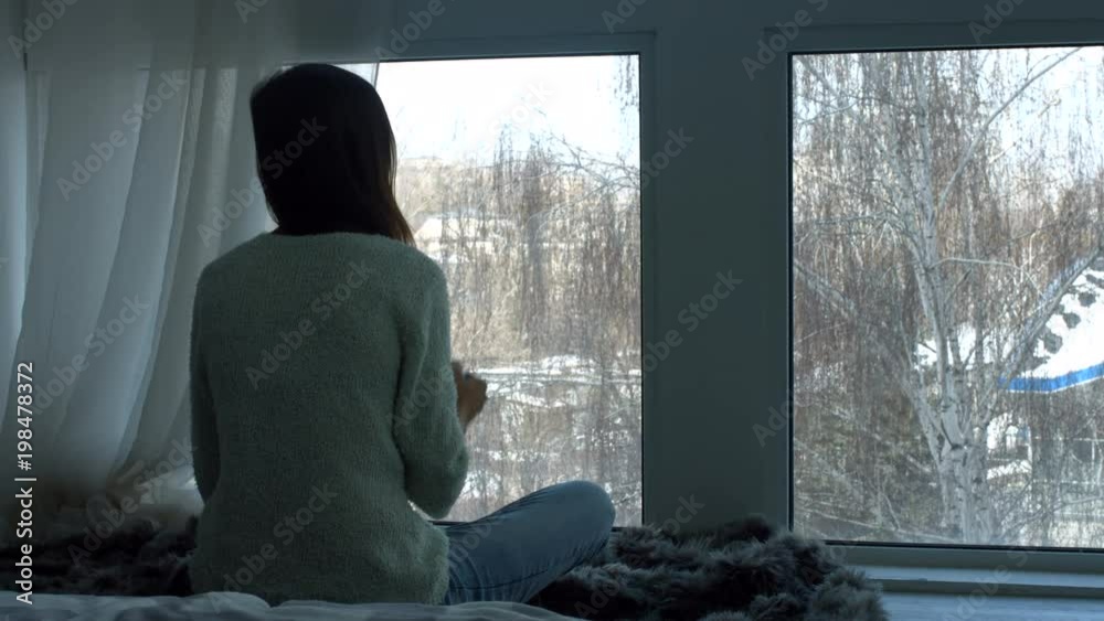 Thoughtful young woman with cup of coffee looking through the window while sitting cross legged on the floor in domestic room. Rear view. Elegant female enjoying mug of coffee in loft apartment.