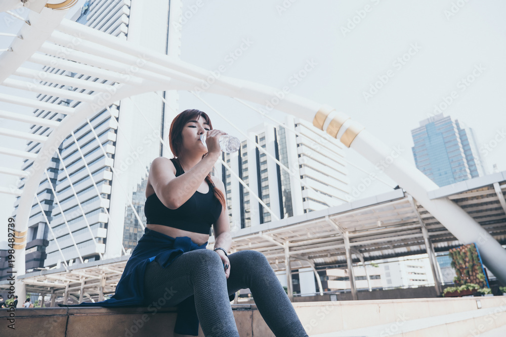 Young Asian woman is relaxing and drinking water after jogging in city. Young Asian woman is relaxing and drinking water after jogging in city.