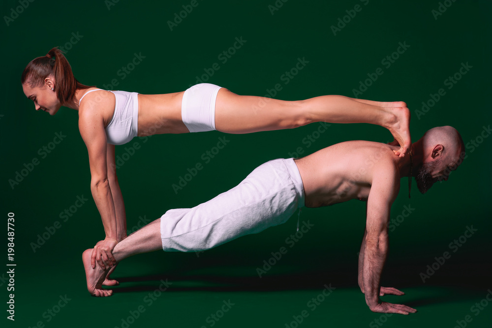 Beautiful sporty woman and man in white clothes doing yoga asanas together indoor on green background