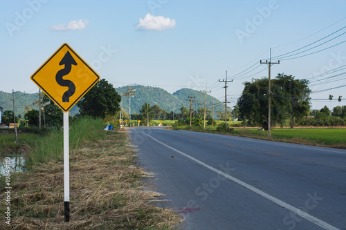 A yellow and black road sign with a winding road symbol stands on the side of a paved road. The road leads towards a rural landscape with green fields and hills in the distance.