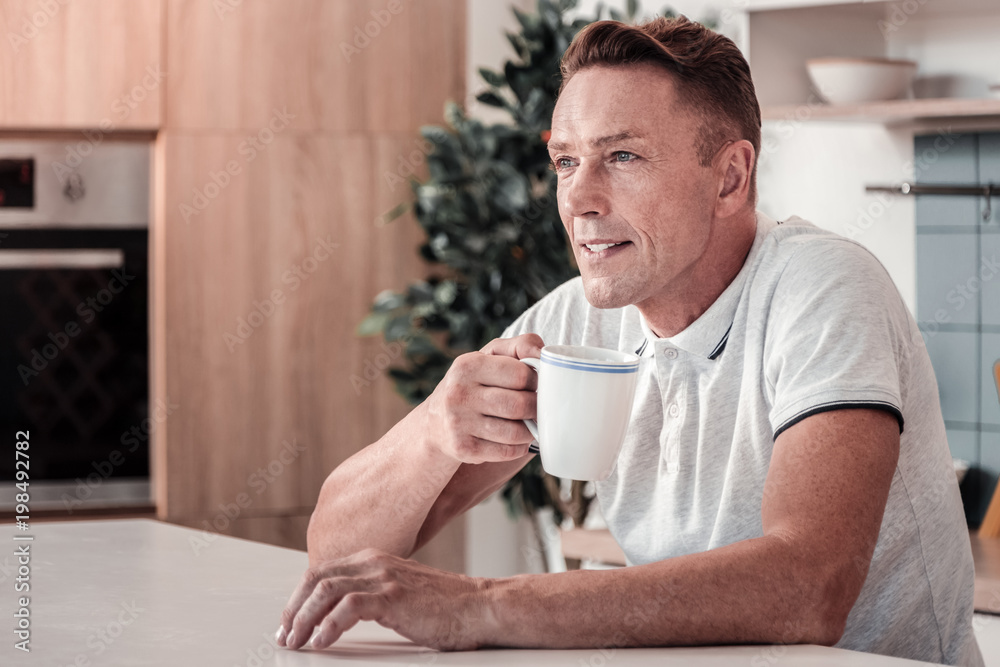 Thoughtful mood. Delighted man keeping smile on his face and leaning elbows on table while creating plan