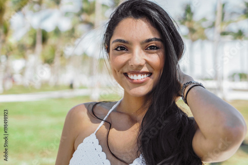 Portrait of beautiful ethnic brunette with long hair smiling at camera on background of tropical park.