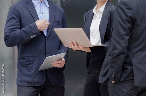 group of business people in suit talking and reading information about finance news in laptop computer together standing in modern city, network technology, internet, successful, teamwork concept