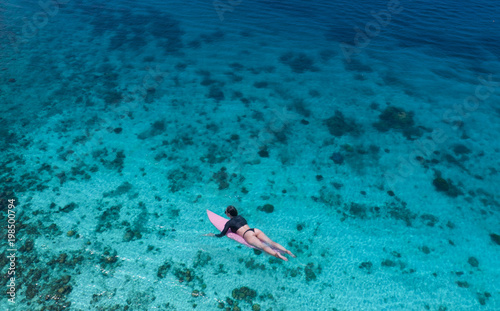 Aerial drone view of beautiful young woman in swimsuit and sunglasses with pink surfboard enjoying her holiday in amazing clear sea water