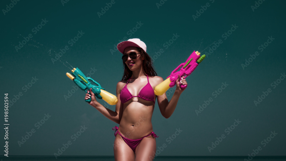Beautiful smiling girl in pink bikini, cap and blue sunglasses splashing water with water guns on the beach over sea and sky background Beautiful smiling girl in pink bikini, cap and blue sunglasses splashing water with water guns on the beach over sea and sky background