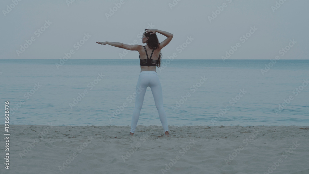 Back view of beautiful young girl exercising in the morning on the sandy beach over sea and sky background