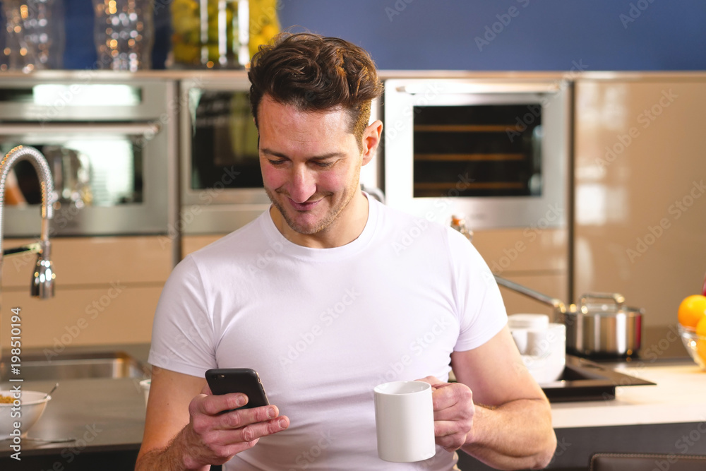A man in the kitchen while having breakfast and sipping tea or milk in the cup sends a message or calls with the tablet and smiles. Concept of: social network, message, technology. A man in the kitchen while having breakfast and sipping tea or milk in the cup sends a message or calls with the tablet and smiles. Concept of: social network, message, technology.