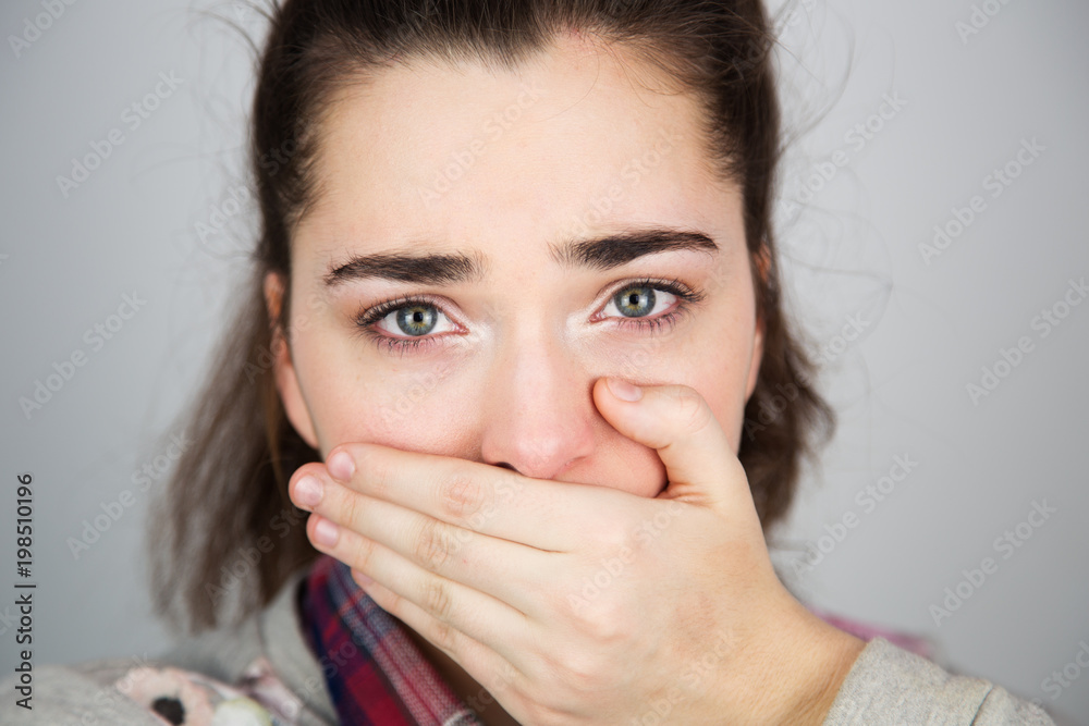 A portrait of a beautiful young caucasian girl with her hand covering mouth on a gray background. Being sad, scared, afraid, abused, hopeless, discouraged. A portrait of a beautiful young caucasian girl with her hand covering mouth on a gray background. Being sad, scared, afraid, abused, hopeless, discouraged.