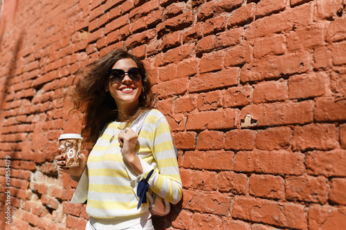 young beautiful girl is walking in summer on a sunny day. he drinks coffee, laughs, writes sms in telephoning or on the Internet, does selfie. Posing on a red brick wall background