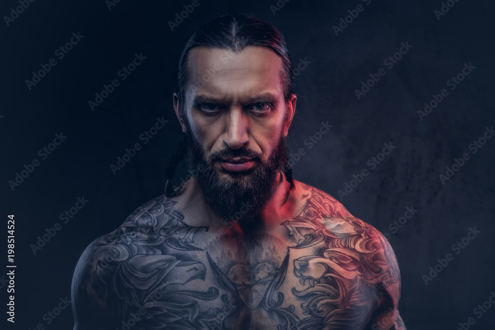 Close-up portrait of a muscular bearded tattoed male with a stylish haircut, isolated on a dark background. Close-up portrait of a muscular bearded tattoed male with a stylish haircut, isolated on a dark background.