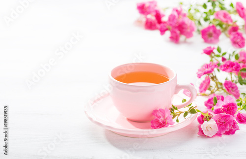 Cup of tea and branch of small pink roses on white rustic table.