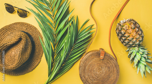 Colorful summer female fashion outfit flat-lay. Straw sunhat, bamboo bag, sunglasses, palm branches and pineapple over yellow background, top view, wide composition. Summer vacation fashion concept