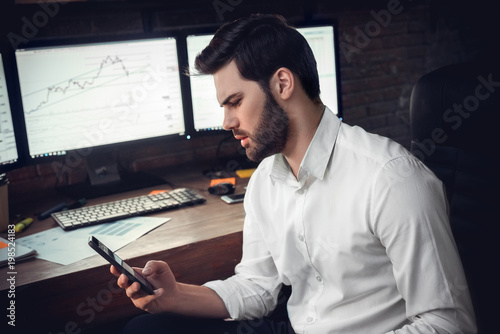 Young male trader at office work concept sitting holding smartphone confused photo