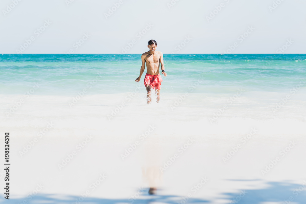 Young man on beautiful sunny white sand beach on Ko Samet, Thailand: Asian man in red swimming trunks walks from turquoise sea waters to the wide empty shore, happy & wet after swim. Wide copy space Young man on beautiful sunny white sand beach on Ko Samet, Thailand: Asian man in red swimming trunks walks from turquoise sea waters to the wide empty shore, happy & wet after swim. Wide copy space