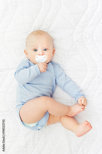 Cute baby boy with pacifier in blue onesie lying on bed