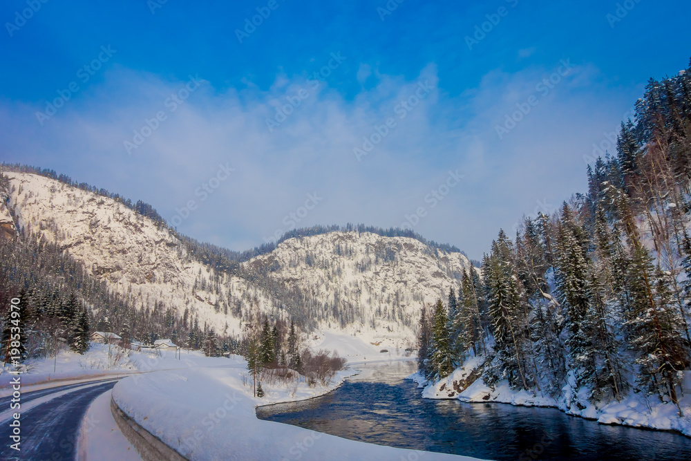 Outdoor view of winter road covered with heavy snow and ice in the forest and small river at one side of the road