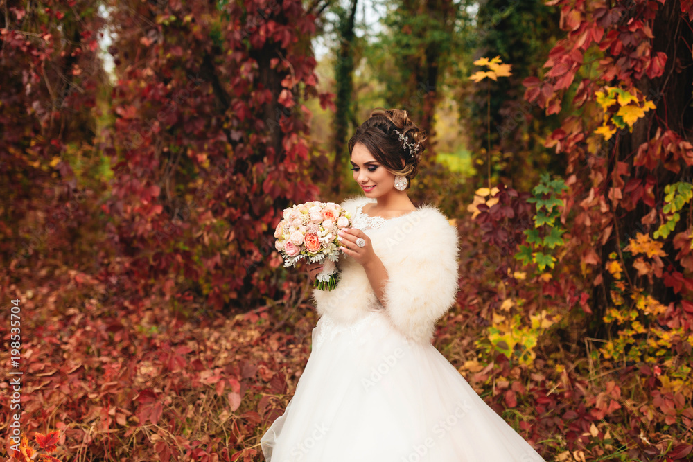 dreamy bride in a wedding dress standing in an autumn forest and looking at her bouquet