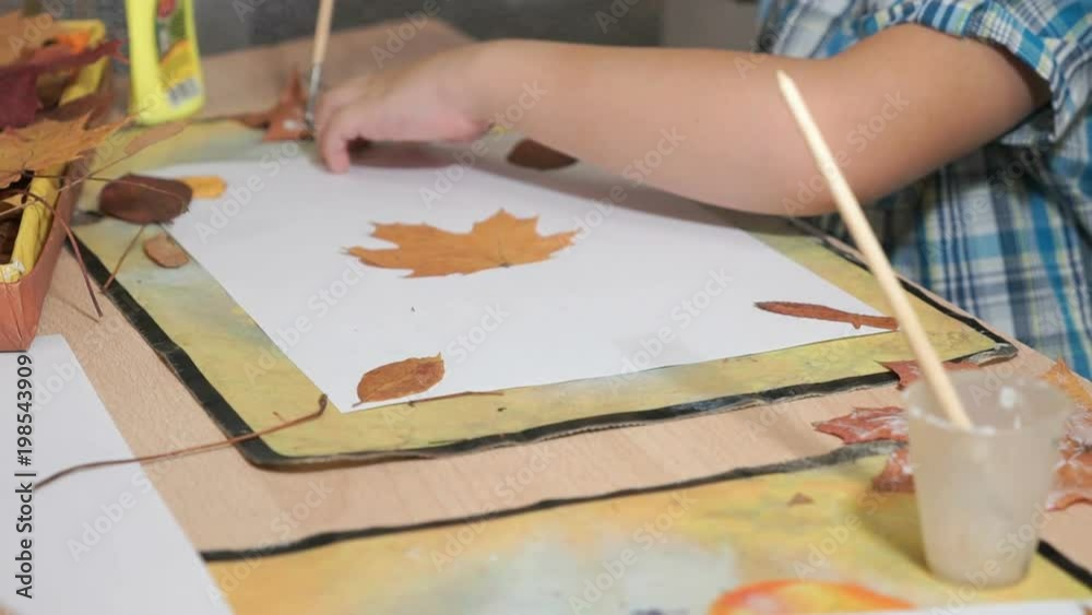 Unknown little child sitting on the table glues dry maple leaves on a white sheet. Beautiful herbarium from leaves of tree. Manual work. Child development in kindergarten Unknown little child sitting on the table glues dry maple leaves on a white sheet. Beautiful herbarium from leaves of tree. Manual work. Child development in kindergarten