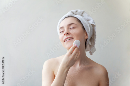 Beauty portrait of a smiling brunette woman in towel on head with soft healthy skin removing make up with cotton pad isolated over white background. Skincare cleansing spa relax concept