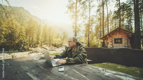 Adult partly bald hunter is sitting outdoors near his shack at the wooden table and working on his laptop; aged businessman in overalls is working remotely on the netbook during his vacations photo