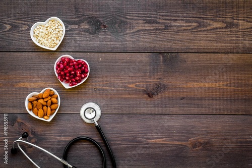 Proper nutrition for pathients with heart disease. Cholesterol reduce diet. Oatmeal, pomegranate, almond in heart shaped bowl near stethoscope on dark wooden background top view copy space